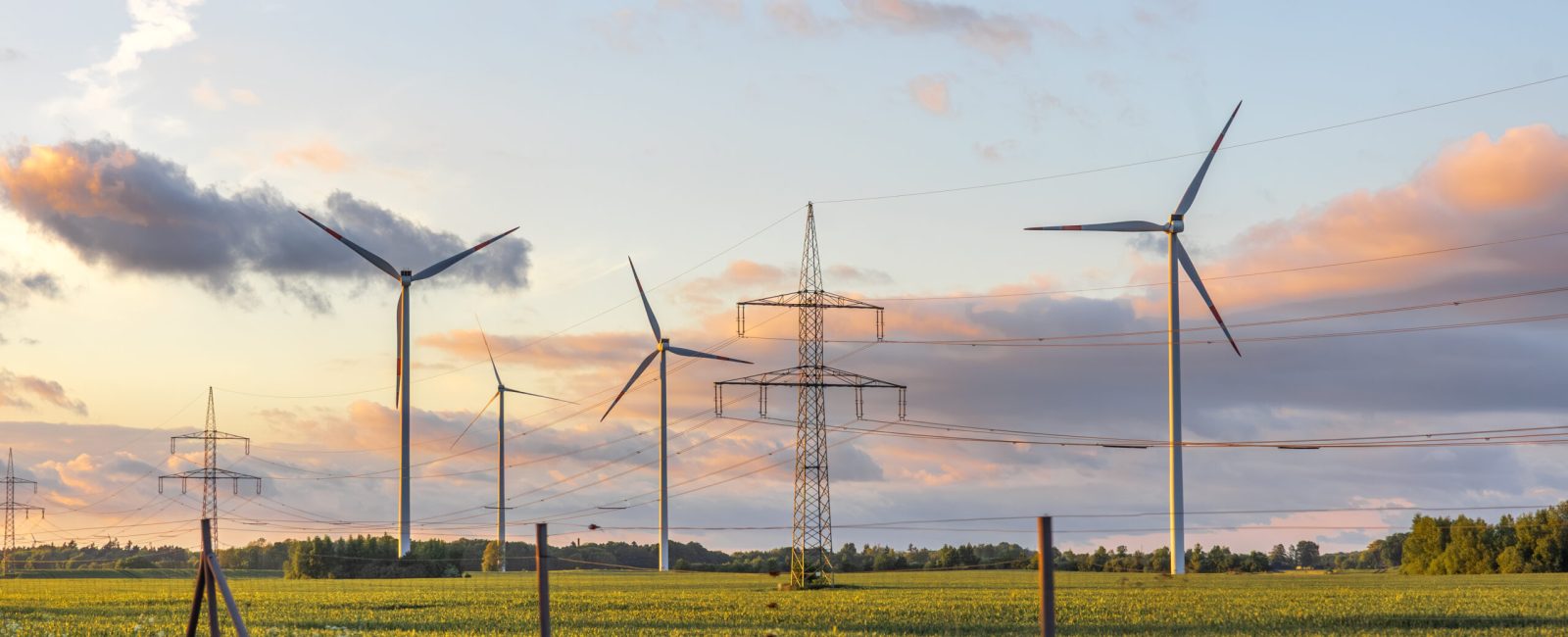 Wind turbines stand alongside power lines in a rural field under pastel sunset skies, blending renewable energy and modern infrastructure.